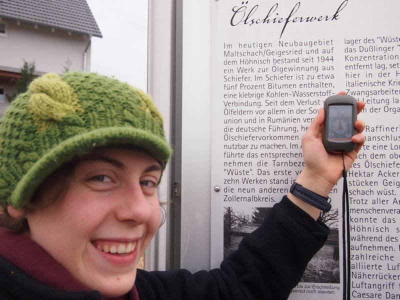 Junge Frau hält ein Geocache-Gerät in der Hand, sie steht vor einer Infotafel mit der Überschrift "Ölschieferwerk"