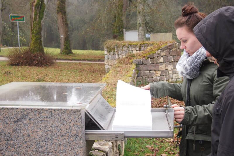 Zwei Personen blättern in einem in einem Steinblock mit Schublade liegenden Buch an einem Ort mit Teil einer Mauer im Freien