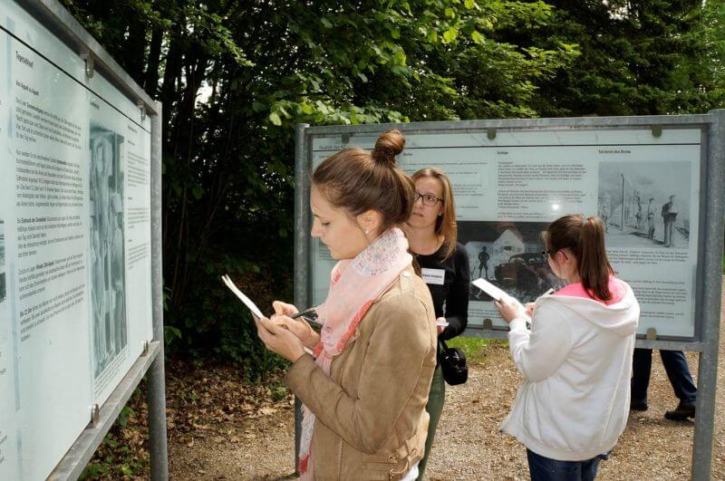 Mehrere junge Menschen stehen bei Gedenktafeln im Freien und machen Notizen