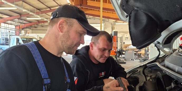 Bildausschnitt: Zwei Personen in Arbeitskleidung in einer Werkstatt an einer geöffneten Motorhaube tät