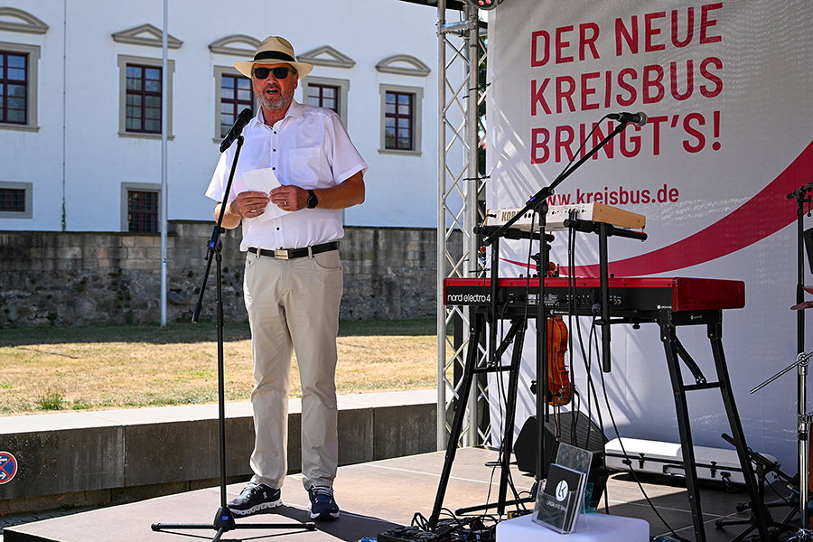 Landrat Joachim Walter spricht am Mikrofon auf der Bühne, daneben ein Keyboard und dahinter das Plakat "Der neue Kreisbus bringt's!"
