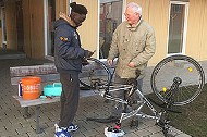 Ein Fahrrad auf den Kopf gestellt, ein junger Mann mit schwarzer Hautfarbe und ein älterer Mann mit weißer Hautfarbe arbeiten daran.