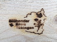 Holz mit eingebranntem Logo: Beschriftung Blühender Kreis Tübingen und Umriss des Landreises mit Blume und Schmetterling