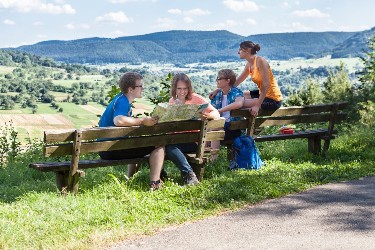 Vier junge Menschen sitzen an einem Aussichtspunkt auf zwei Bänken, sie schauen zu den Bergen der Schwäbischen Alb und in eine Karte