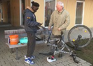 Ein Fahrrad auf den Kopf gestellt, ein jüngerer Mann mit schwarzer Hautfarbe und ein älterer Mann mit weißer Hautfarbe bei der Reparatur