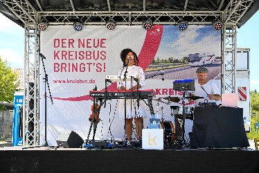 Eine Musikerin steht und ein Musiker sitzt auf einer Bühne vor einem großen  Plakat mit der Aufschrift "Der neue Kreisbus bringt's!, www.kreisbus.de"