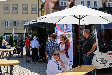 Personen auf einem Platz mit Infotafeln, Sonnenschirmen und Tischen