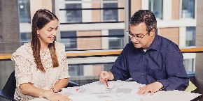 Zwei Personen an einem Tisch, eine Person zeigt erklärend mit dem Bleistift Inhalte auf Arbeitsunterlagen