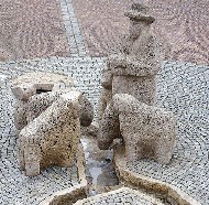 Ebenerdig mit Pflastersteinen und eingefasster Rinne angelegter Brunnen, mit einer Gruppe Steinfiguren: ein Hirte mit  Tiere