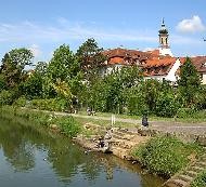 Fluss mit Uferweg, Sitzgelegenheiten und Bepflanzung im Stadtzentrum, dahinter historische Gebäude und Kirchturm