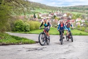 Drei Mountainbike-Fahrer*innen auf einem Weg in Landschaft mit Ortschaft, Link zur Tourismus-Seite tuebinger-umwelten.de